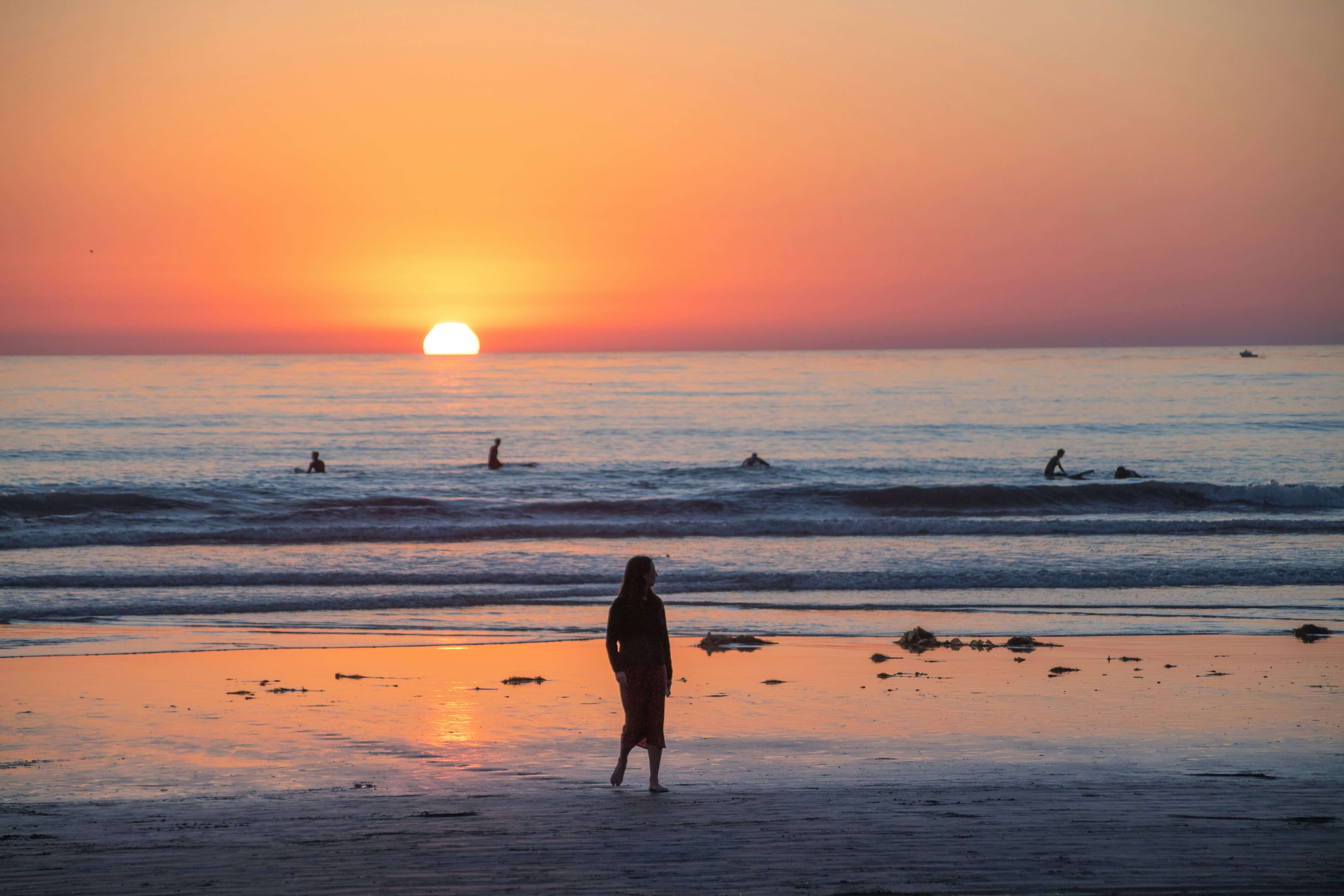 Mulher de costas caminhando na beira do mar ao pôr do sol